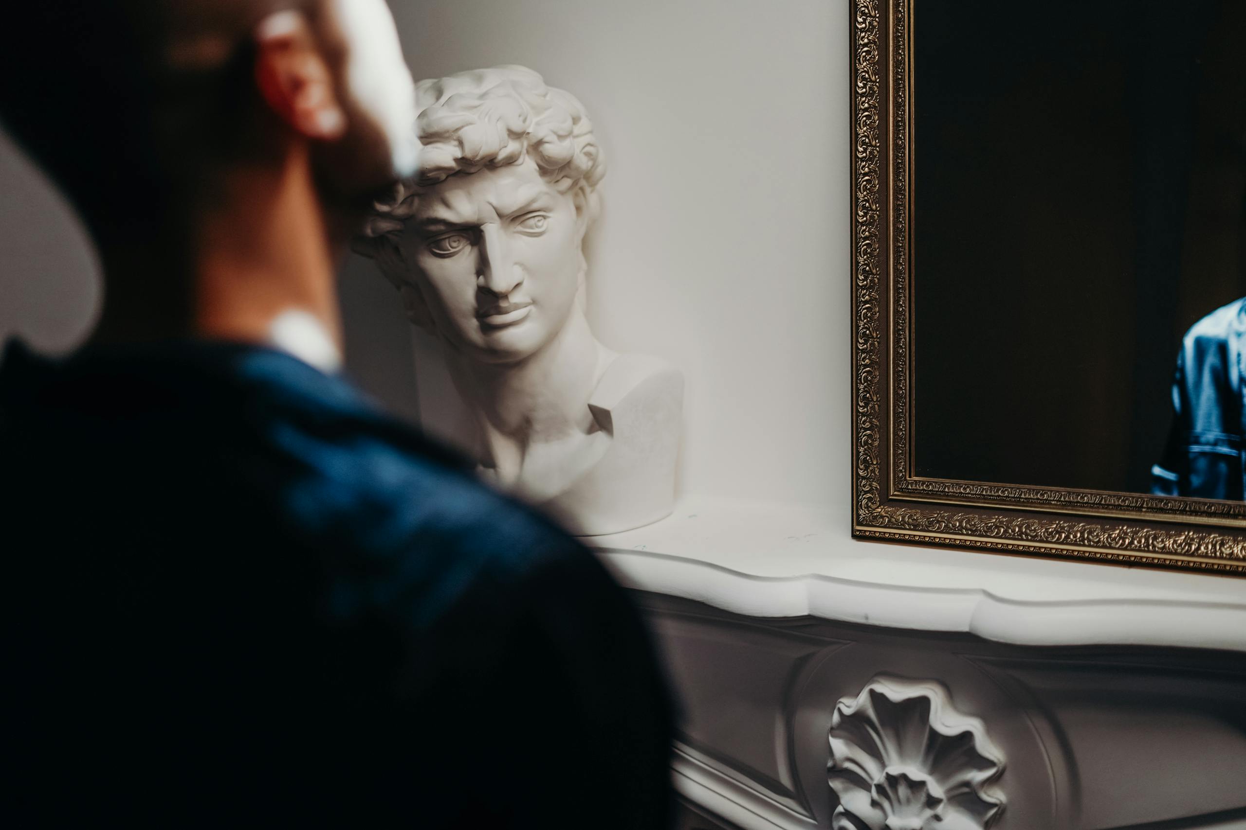 A man gazes into a mirror beside a classic sculpture in a sophisticated room.
